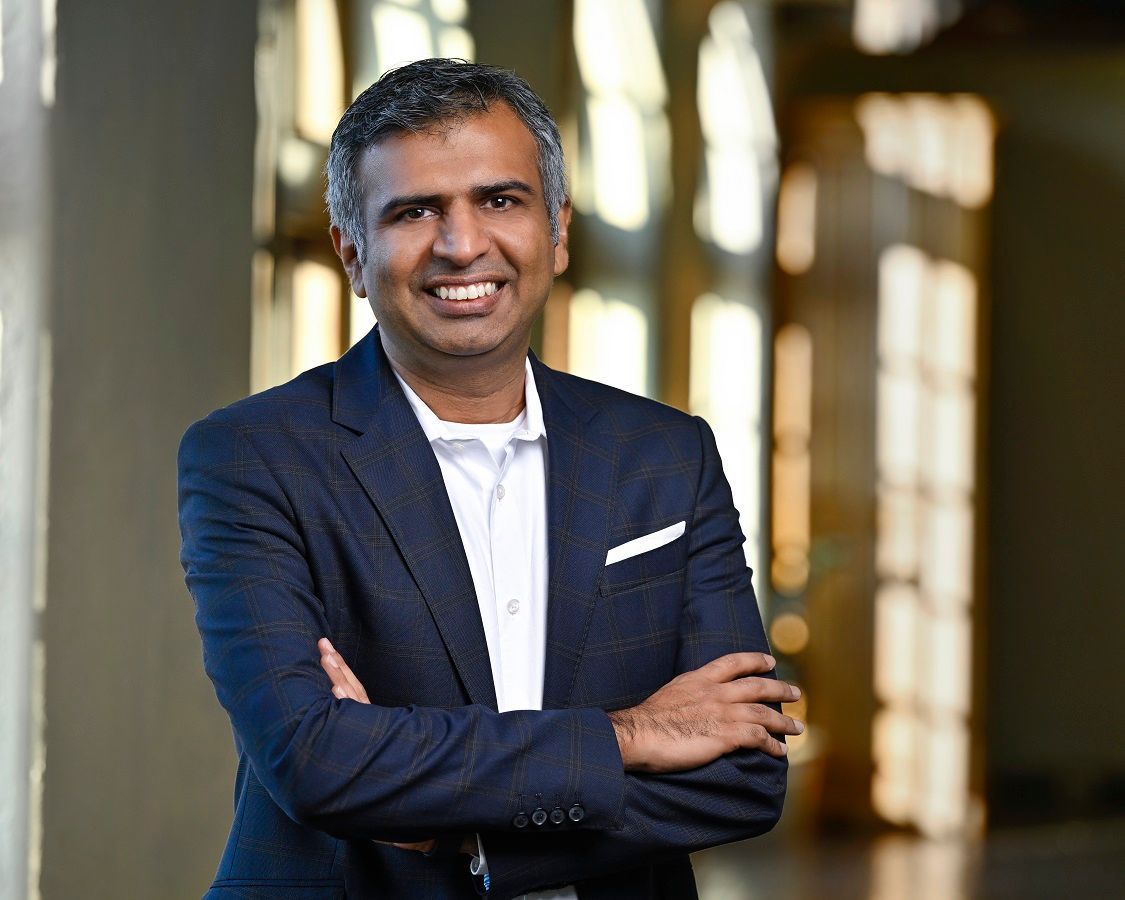 A smiling man in a navy blazer and white collared shirt stands with arms crossed against a blurred indoor backdrop.