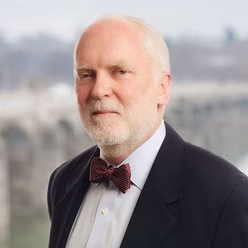 A man with a white beard wearing a dark suit and a patterned bow tie, posed against a blurred outdoor background.