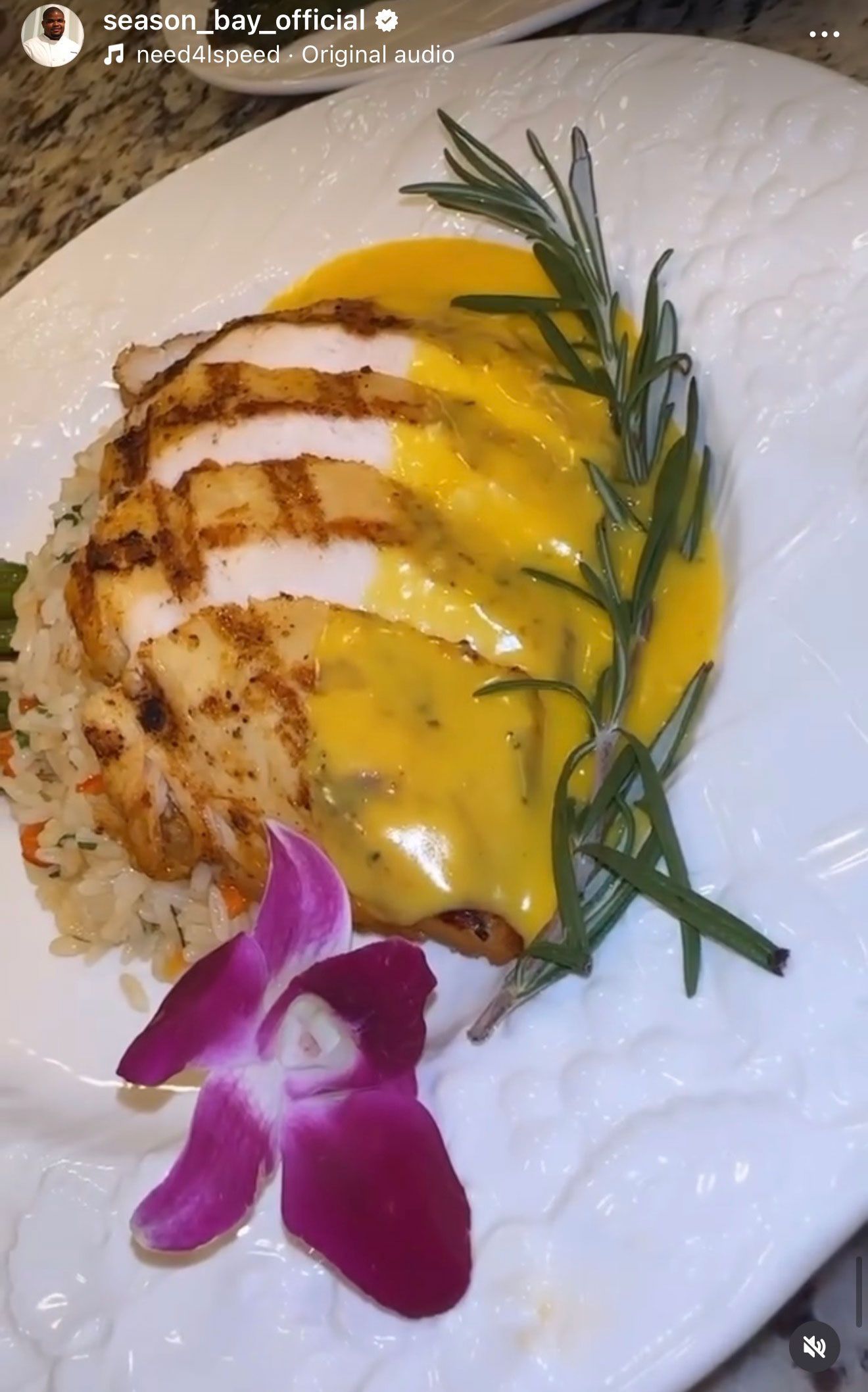Grilled chicken with yellow sauce and rice, garnished with rosemary and a purple orchid on a white plate.