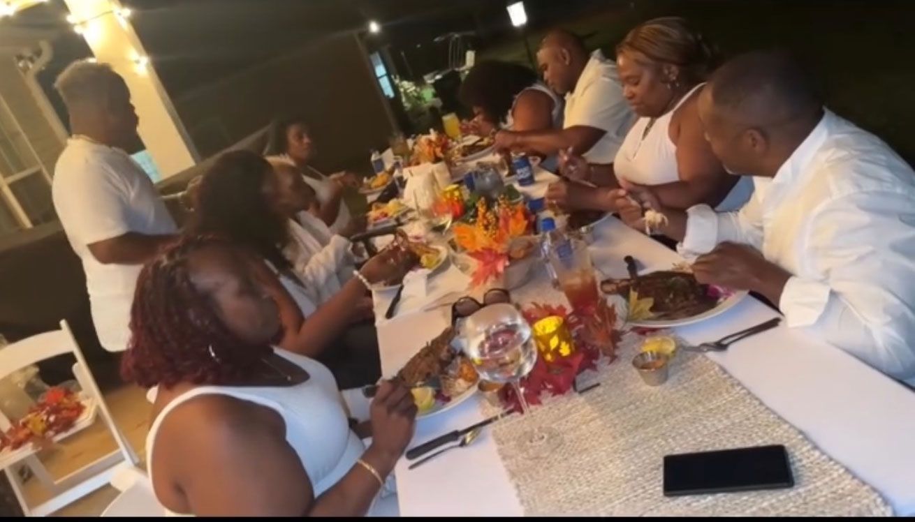 People in white attire seated at a long table with food and decorations, lit by warm lighting.