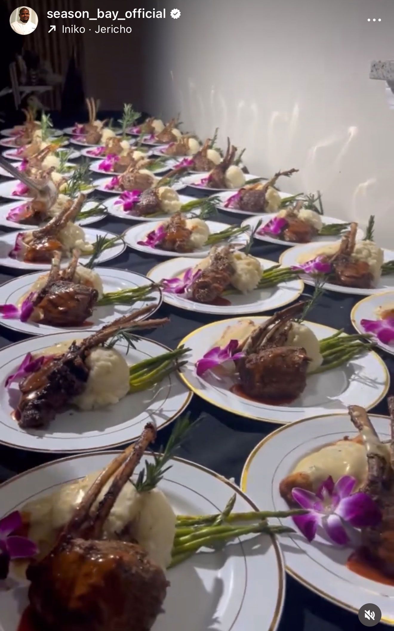 Lamb chops on plates, served with mashed potatoes, asparagus, and purple orchids, arranged in a row on a black tablecloth.