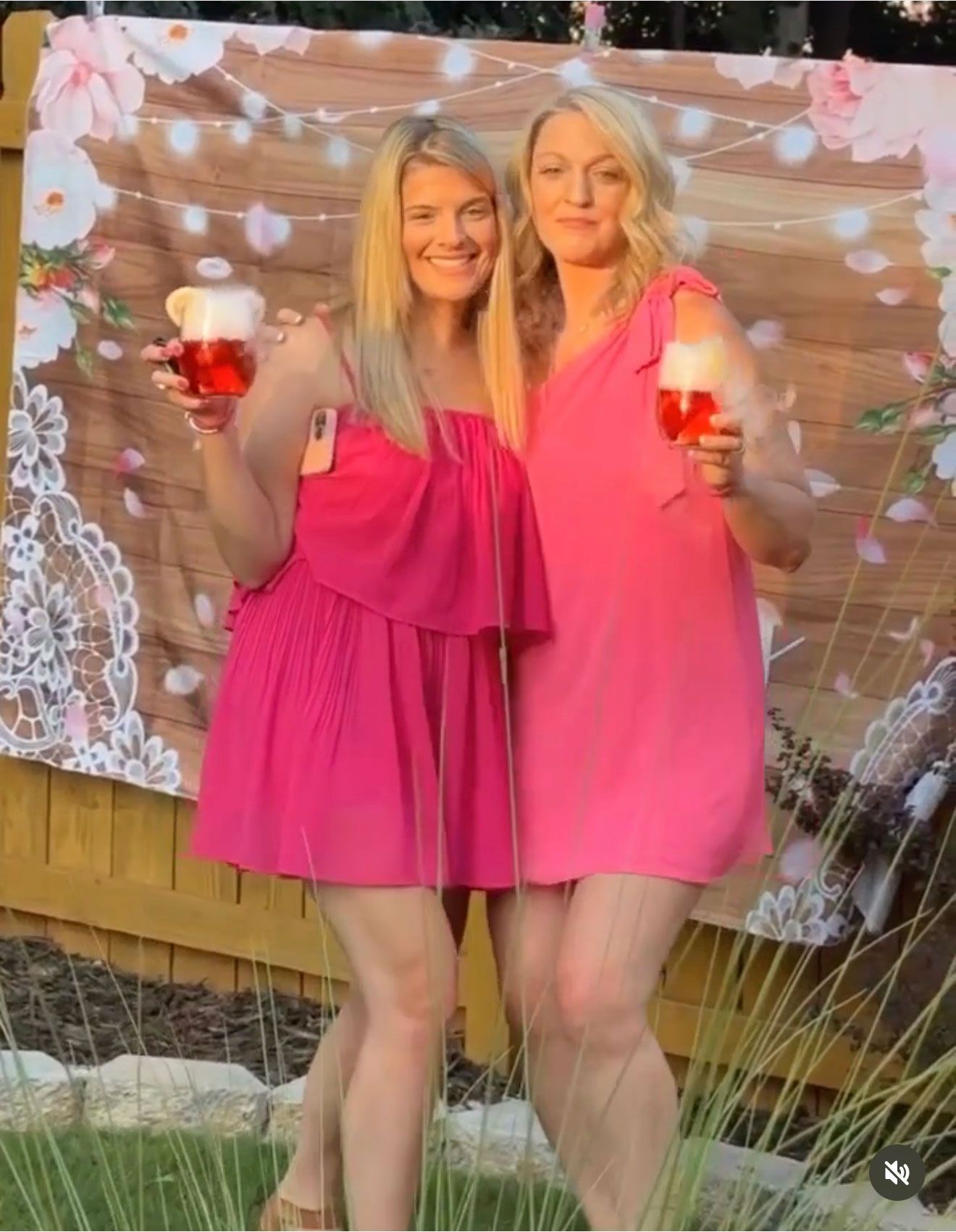 Two women in pink dresses holding drinks, smiling, in front of a backdrop.