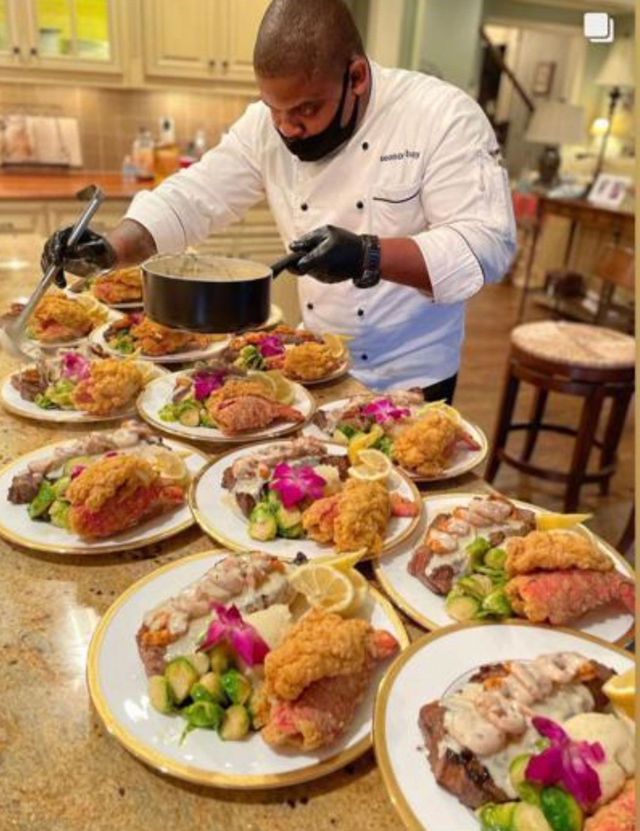 Chef plates multiple dishes with fried fish, shrimp, and vegetables in a well-lit kitchen.