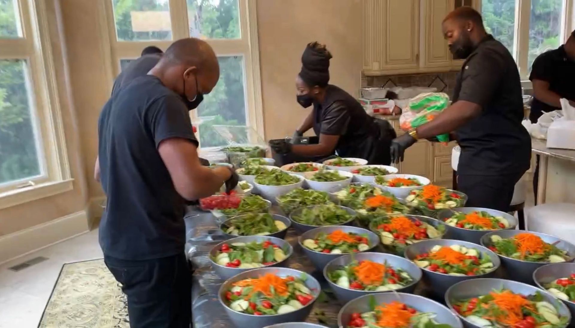 People preparing salads in a kitchen, wearing masks. Bowls of salad on a counter near a window.