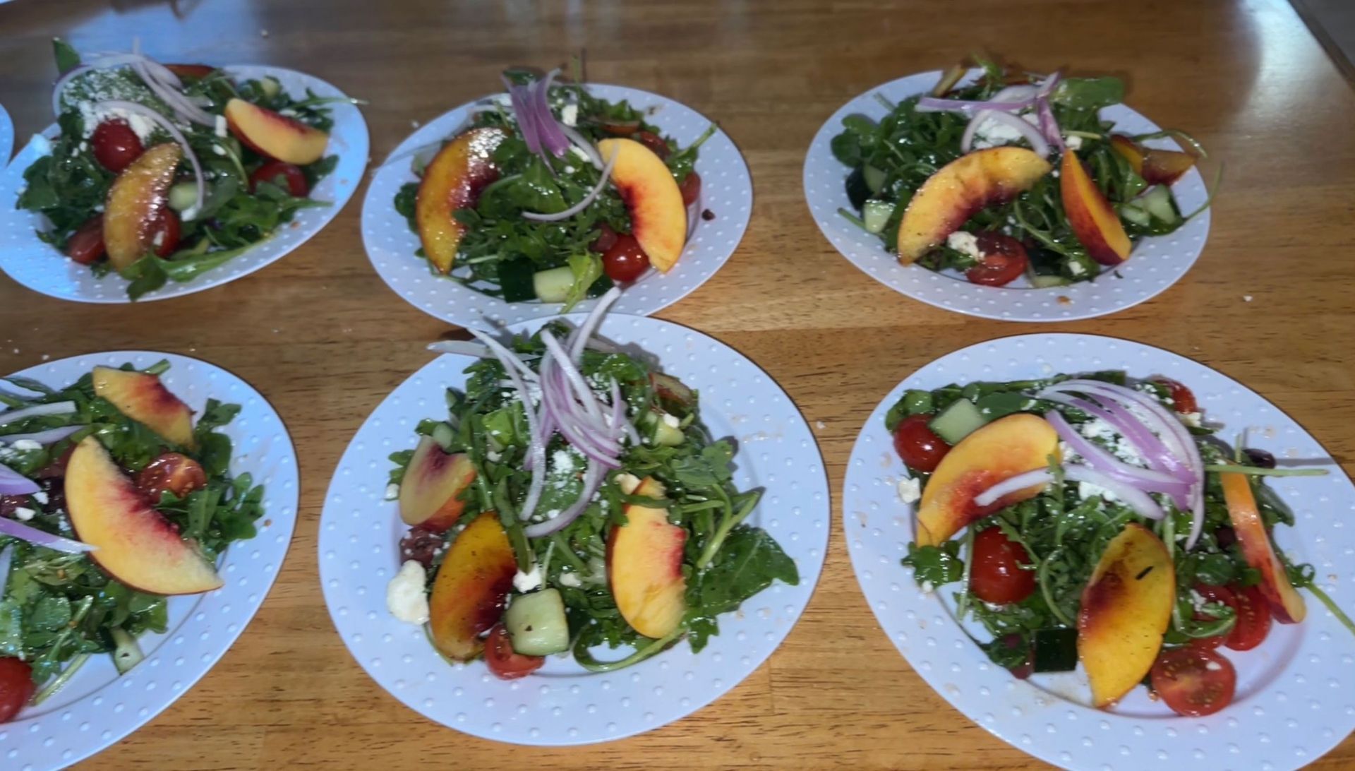 Salads with arugula, peach slices, red onion, tomatoes, and cheese on white plates, arranged on a wooden table.