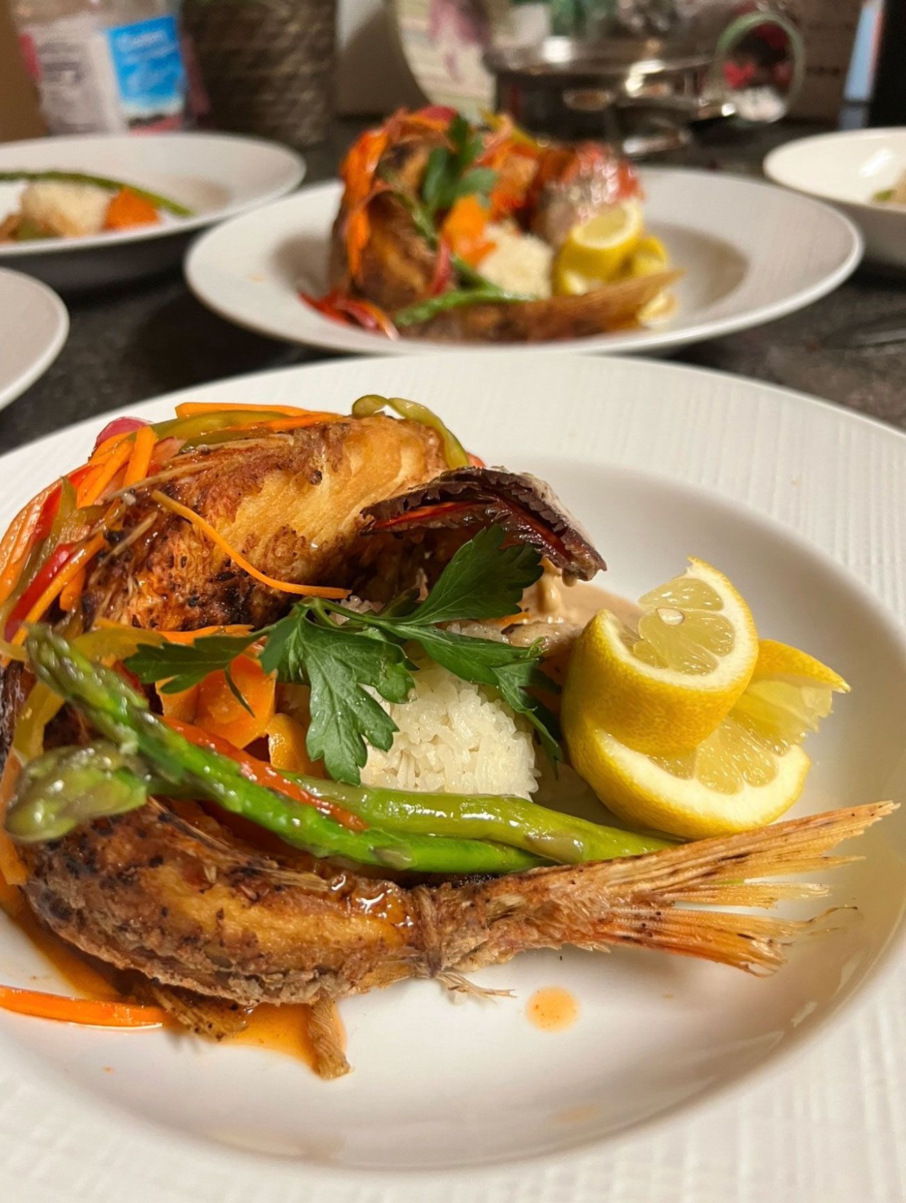 Fried fish on a white plate with rice, vegetables, and lemon slices. Other plates are visible.