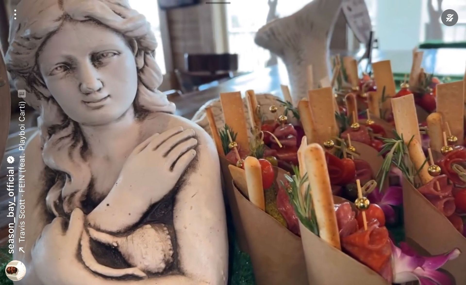 Statue next to food cones: statue of woman with food cones with meats, rosemary, and breadsticks.