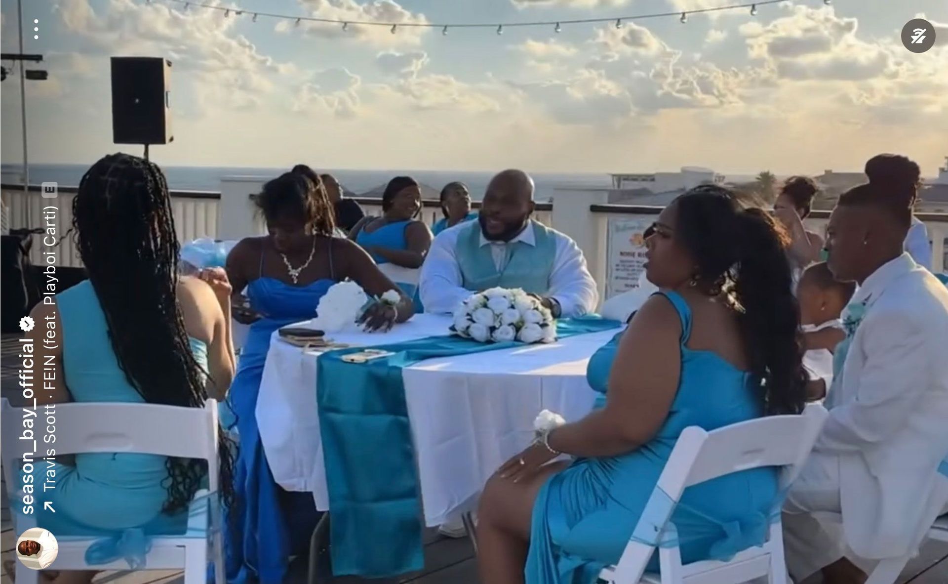 Wedding reception on a rooftop with guests seated around tables draped in blue. Bride and groom at center.