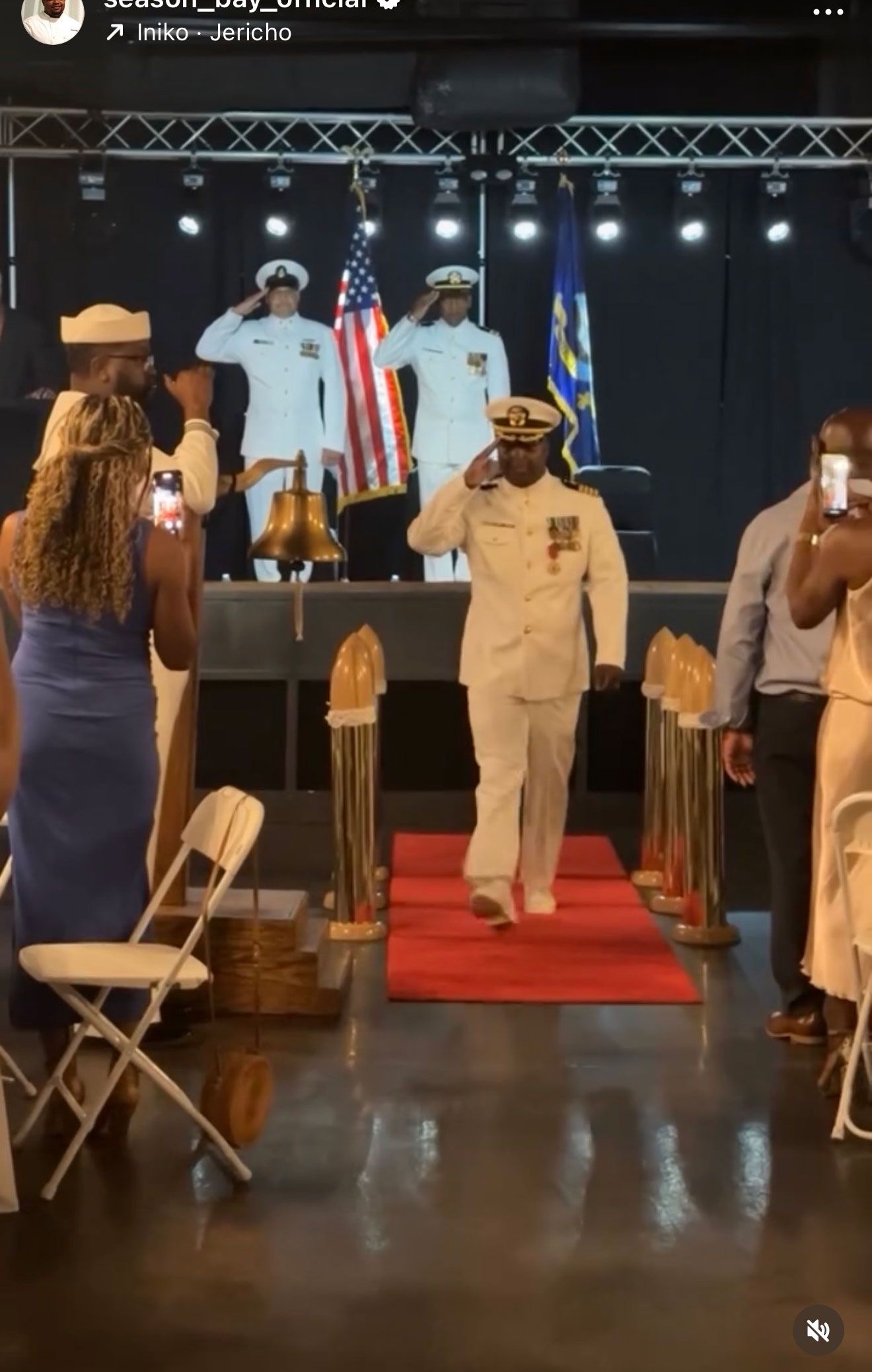 A man in a Navy uniform salutes as he walks a red carpet. Other servicemen and guests watch.
