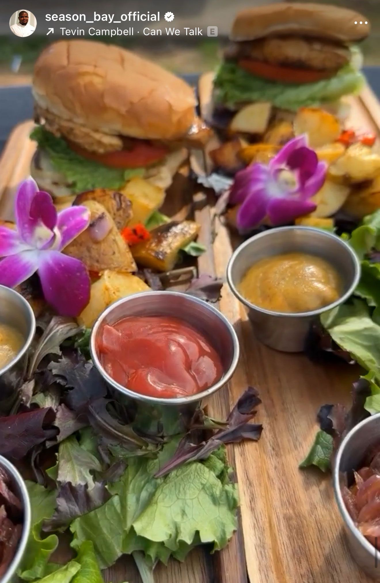 Two burgers with fries and dips, garnished with flowers, served on a wooden board.