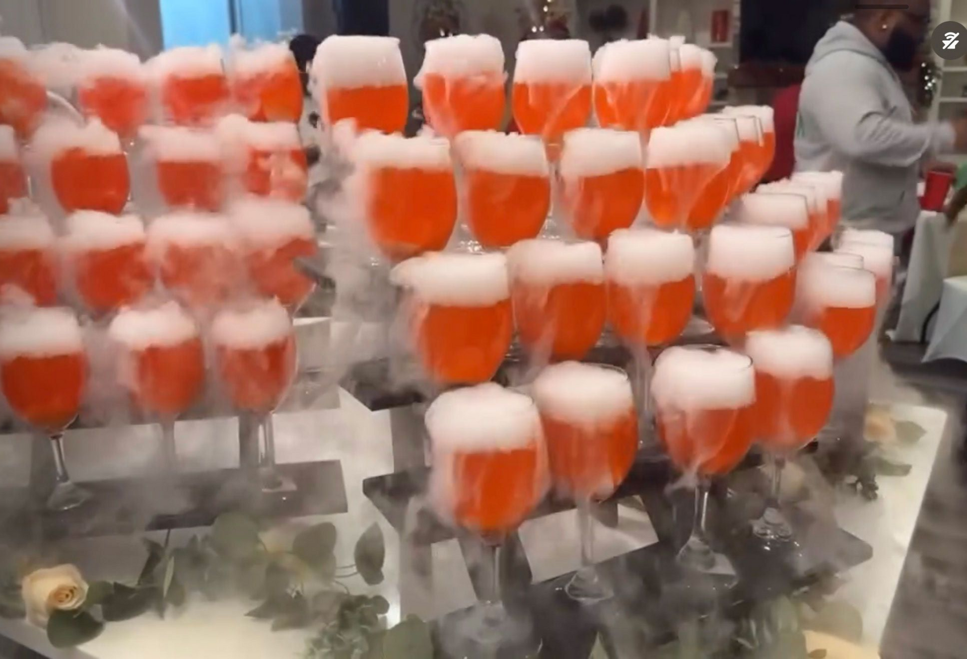 Orange drinks with dry ice, arranged in tiers, emitting vapor. Person in background.