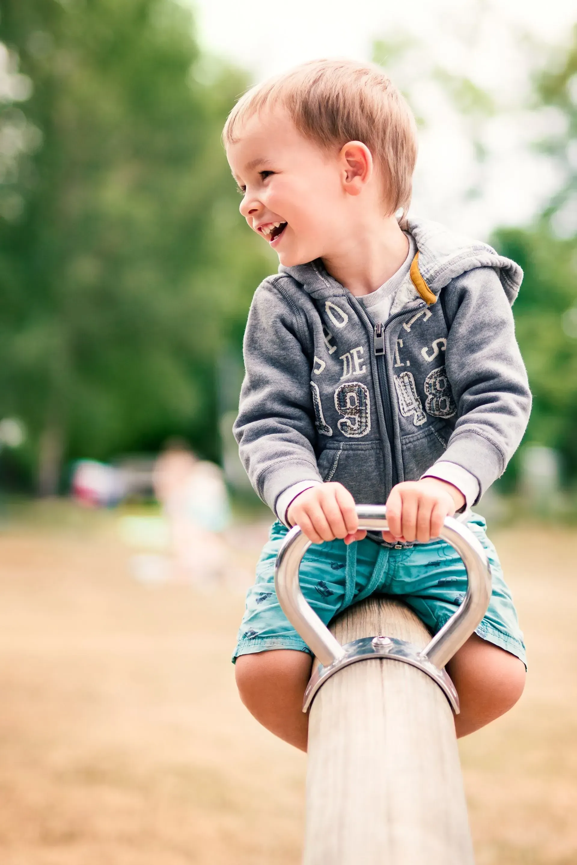 Boy on a seesaw, smiling and laughing, wearing a gray hoodie and teal shorts at a park.