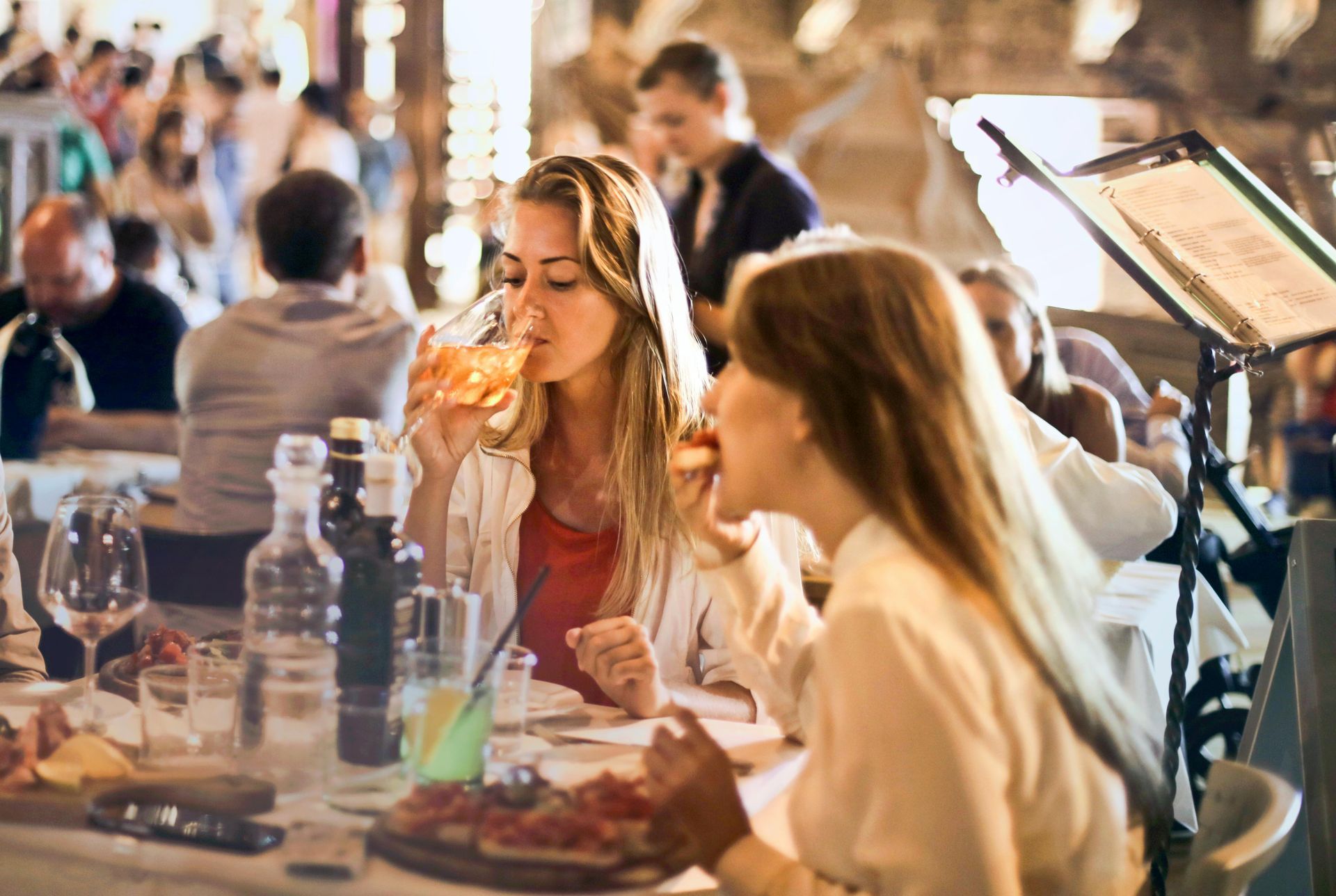 Two women at a restaurant, one drinking, the other eating. Other patrons and server in background.