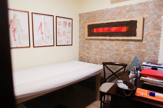 Una sala per trattamenti con un lettino da massaggio, tre tavole anatomiche a parete e una scrivania con un caminetto decorativo.