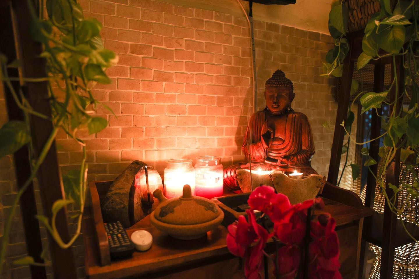 Una statua di Buddha si erge su un altare di legno illuminato da candele, addossato a un muro di mattoni ornato da piante e fiori pendenti.