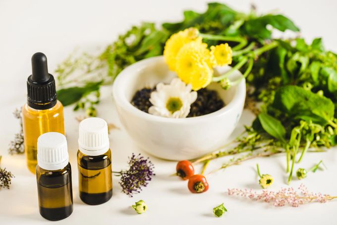 Tre boccette di oli essenziali, un mortaio con fiori gialli e bianchi e varie erbe aromatiche verdi su una superficie bianca.