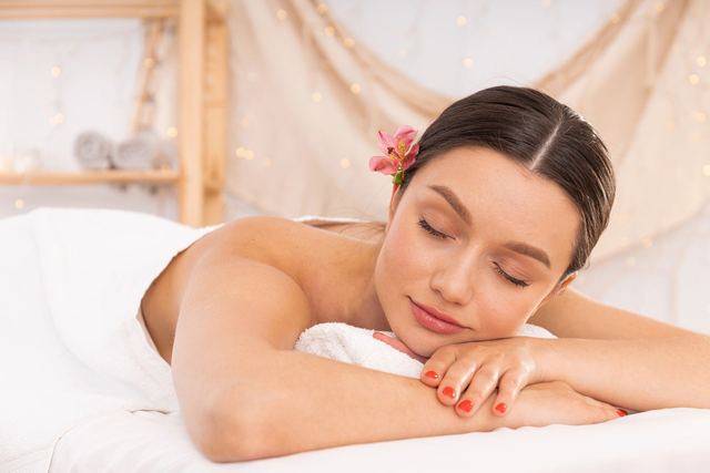 In un ambiente termale, una persona rilassata con un piccolo fiore tra i capelli riposa su un lettino da massaggio ricoperto da un asciugamano bianco.