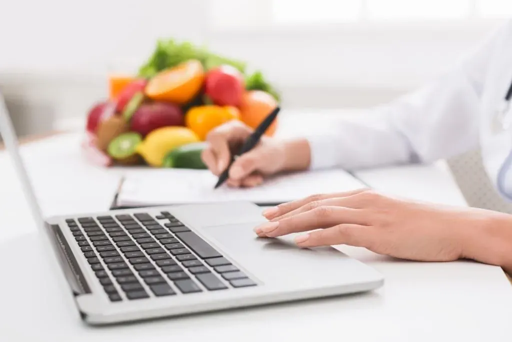 Una persona in camice bianco scrive su un documento accanto a un computer portatile, con sullo sfondo una ciotola sfocata di frutta fresca e colorata.