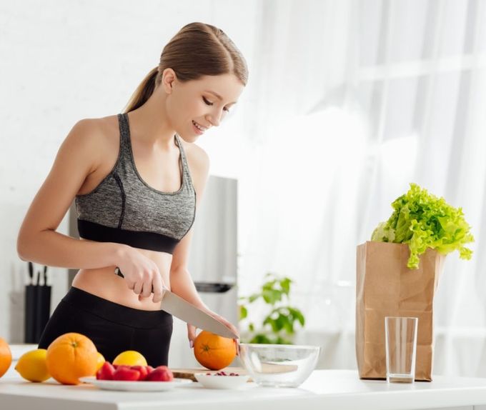 Una persona in abbigliamento sportivo sorride mentre affetta un'arancia su un bancone, accanto ad altra frutta fresca e a un sacchetto di verdure.