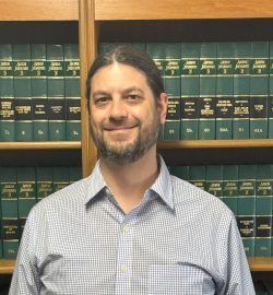 Man with beard smiling in front of a bookshelf with green books. He wears a blue plaid shirt.