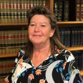 Woman in floral shirt smiles in a library, surrounded by bookshelves.