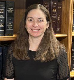 Woman with long brown hair, smiling in front of a bookshelf with legal books. She wears a black top.
