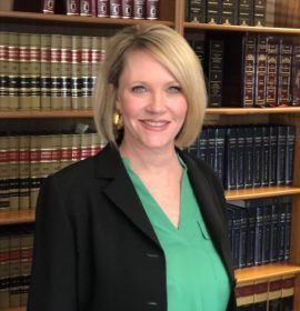 Woman in a blazer and green shirt smiles in front of a bookcase filled with books.
