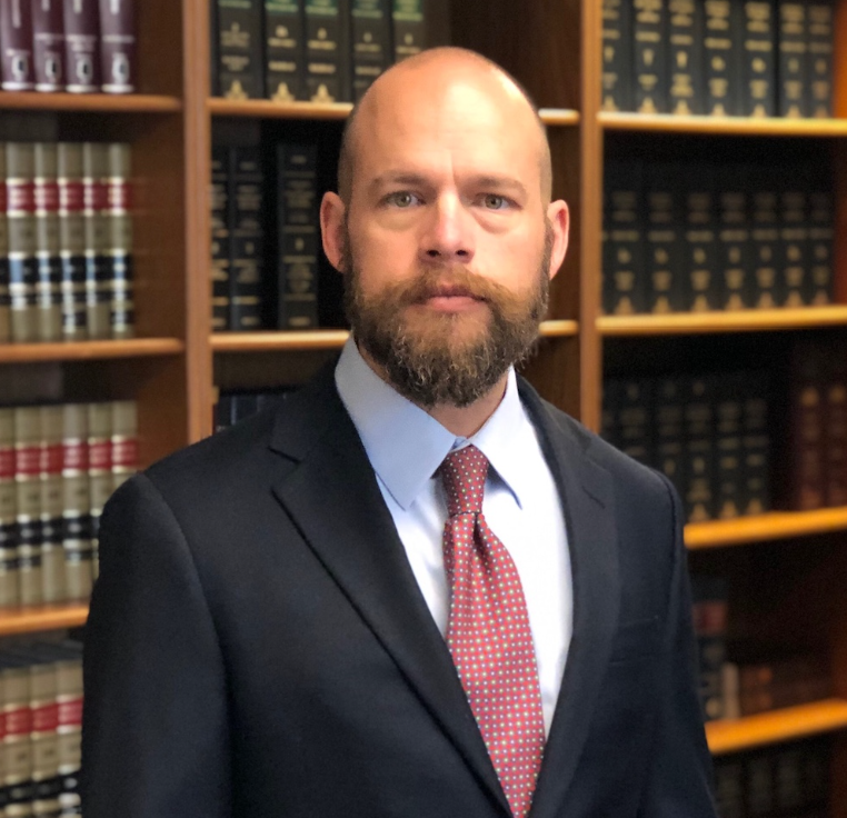 Man in suit and tie with beard, standing in front of a bookshelf filled with books.