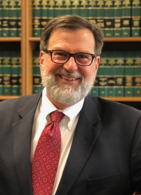 Man in suit and tie, smiling, wearing glasses, in front of a bookshelf.