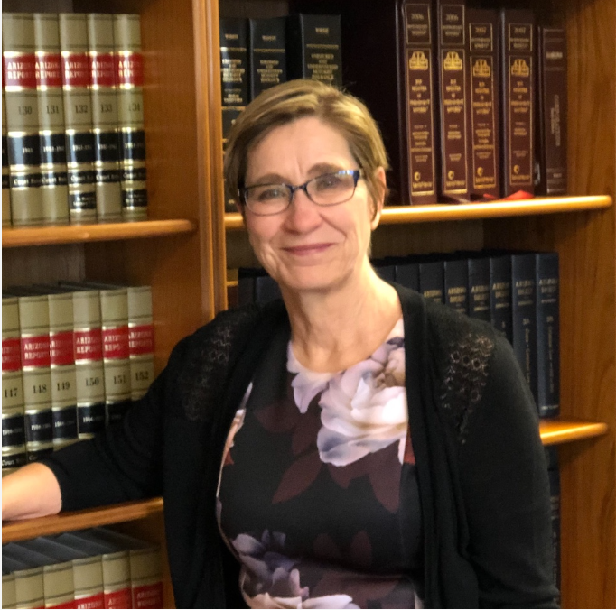 Woman with glasses, stands near a bookcase with law books, wearing a floral dress and black cardigan.