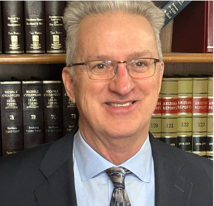 Man in glasses and suit smiling in front of a bookshelf with legal books.