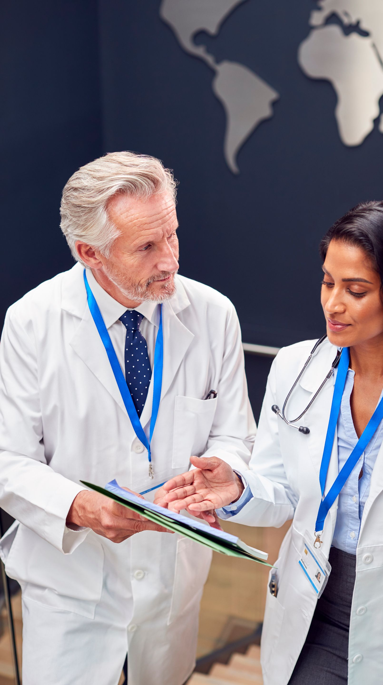 Two doctors in white coats reviewing a tablet; world map in the background.
