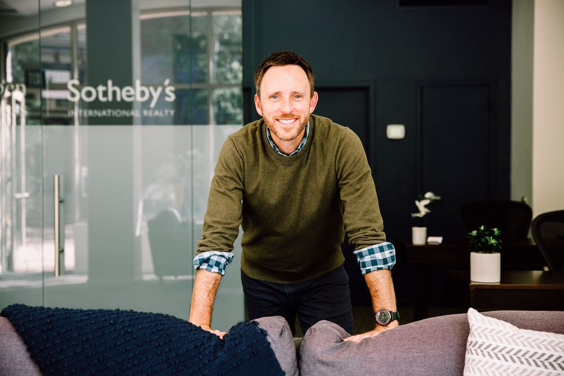 A man in a green sweater smiles while leaning on a couch in a Sotheby's International Realty office.