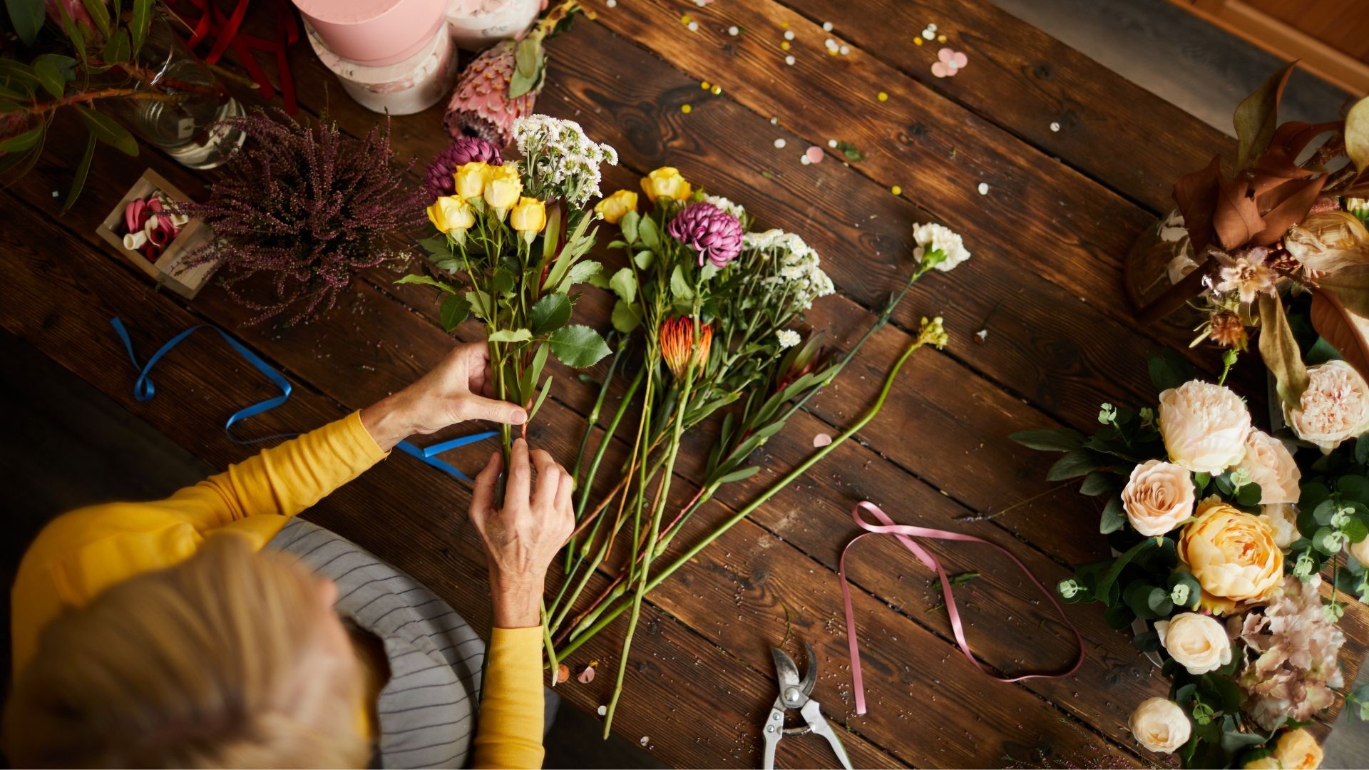 Florist arranging colorful flowers on a wooden table, blue ribbon, scissors, close-up shot.