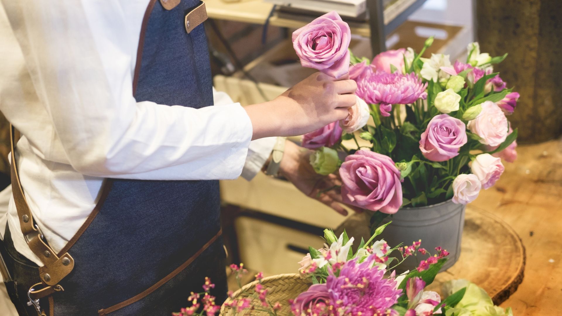 Florist arranging pink roses and other flowers in a gray pot, wearing a blue apron.