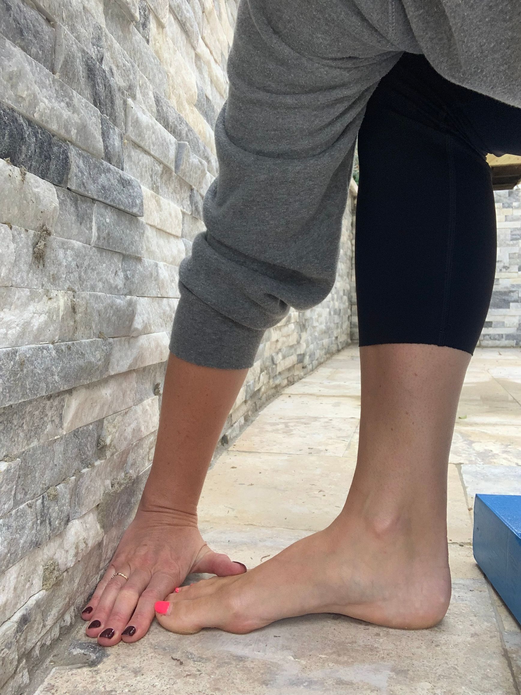 Person stretching foot, hand on wall, leg in black leggings, gray sweater.
