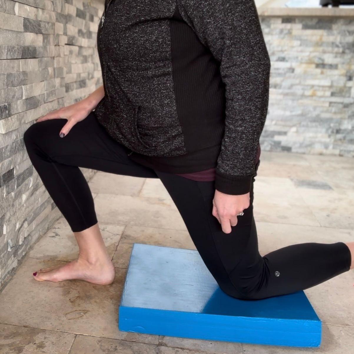 Person stretches their legs, one knee down on a blue mat, by a stone wall.
