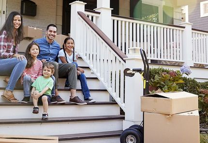 Family sitting on front steps of a house, surrounded by moving boxes; sunshine.