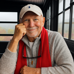 Man in a baseball cap and red scarf smiles, resting his head on his hand. Inside, window behind.