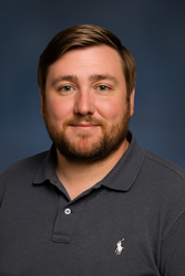 Man with brown hair and beard wearing a gray polo shirt, smiling, against a blue background.