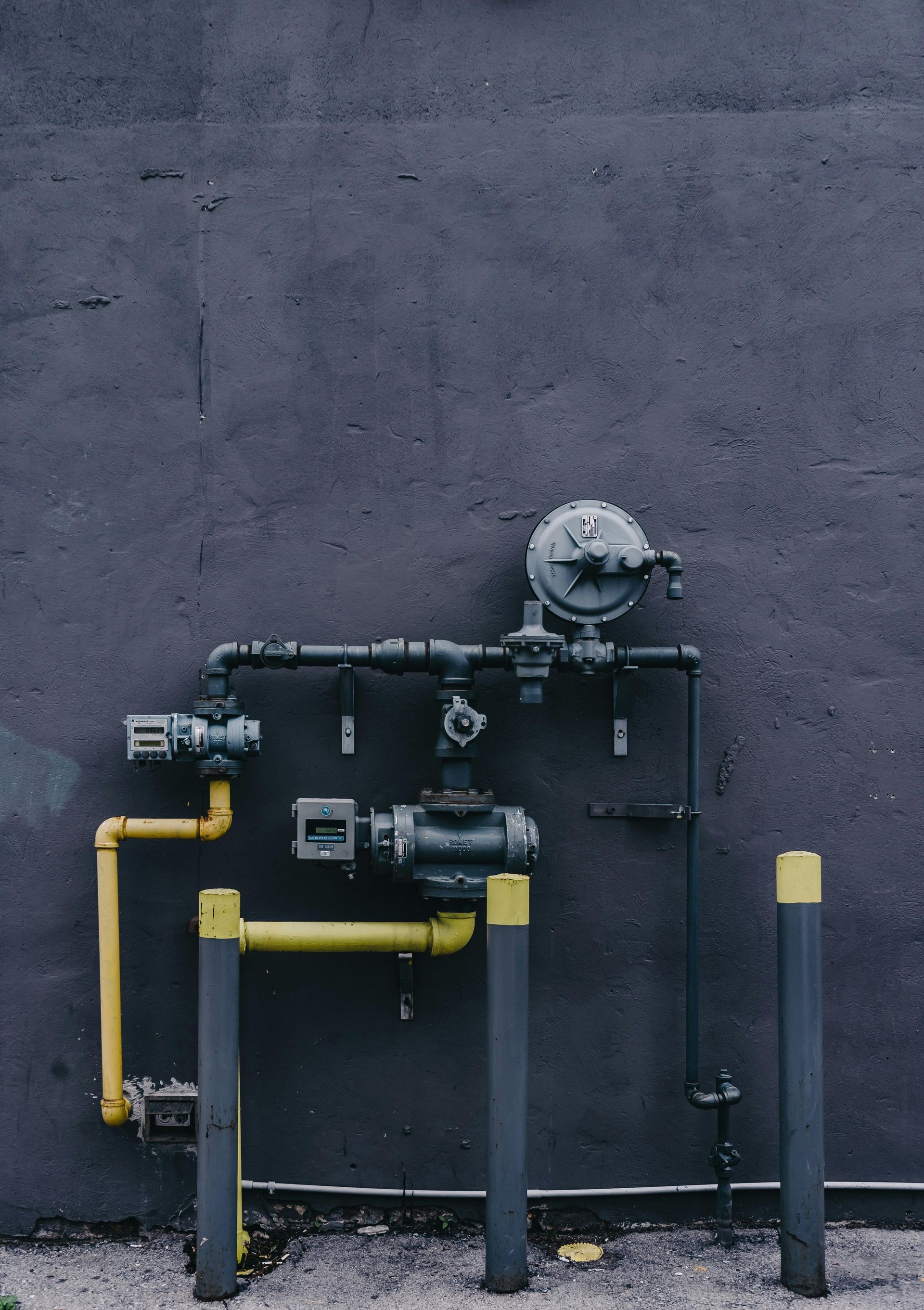 Industrial gas pipes and meters mounted on a dark wall, with yellow piping accents.