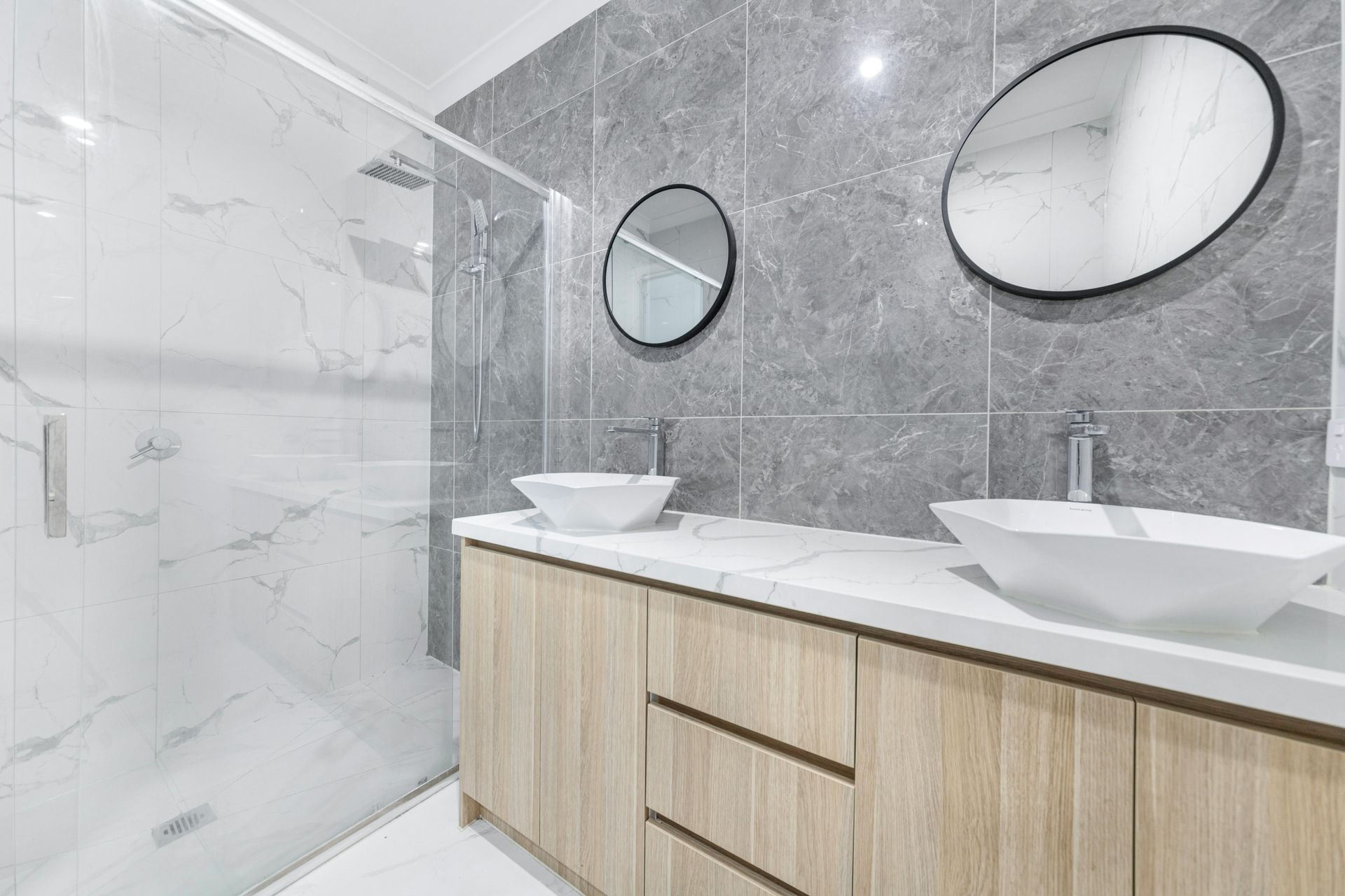 Modern bathroom with double sinks, round mirrors, gray tile walls, and a glass shower