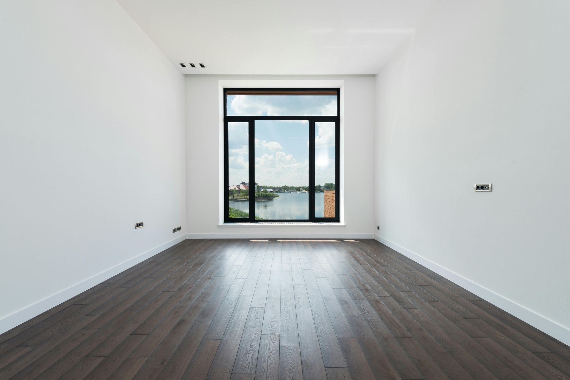 Empty modern room with dark wood floors, white walls, and a large window overlooking greenery