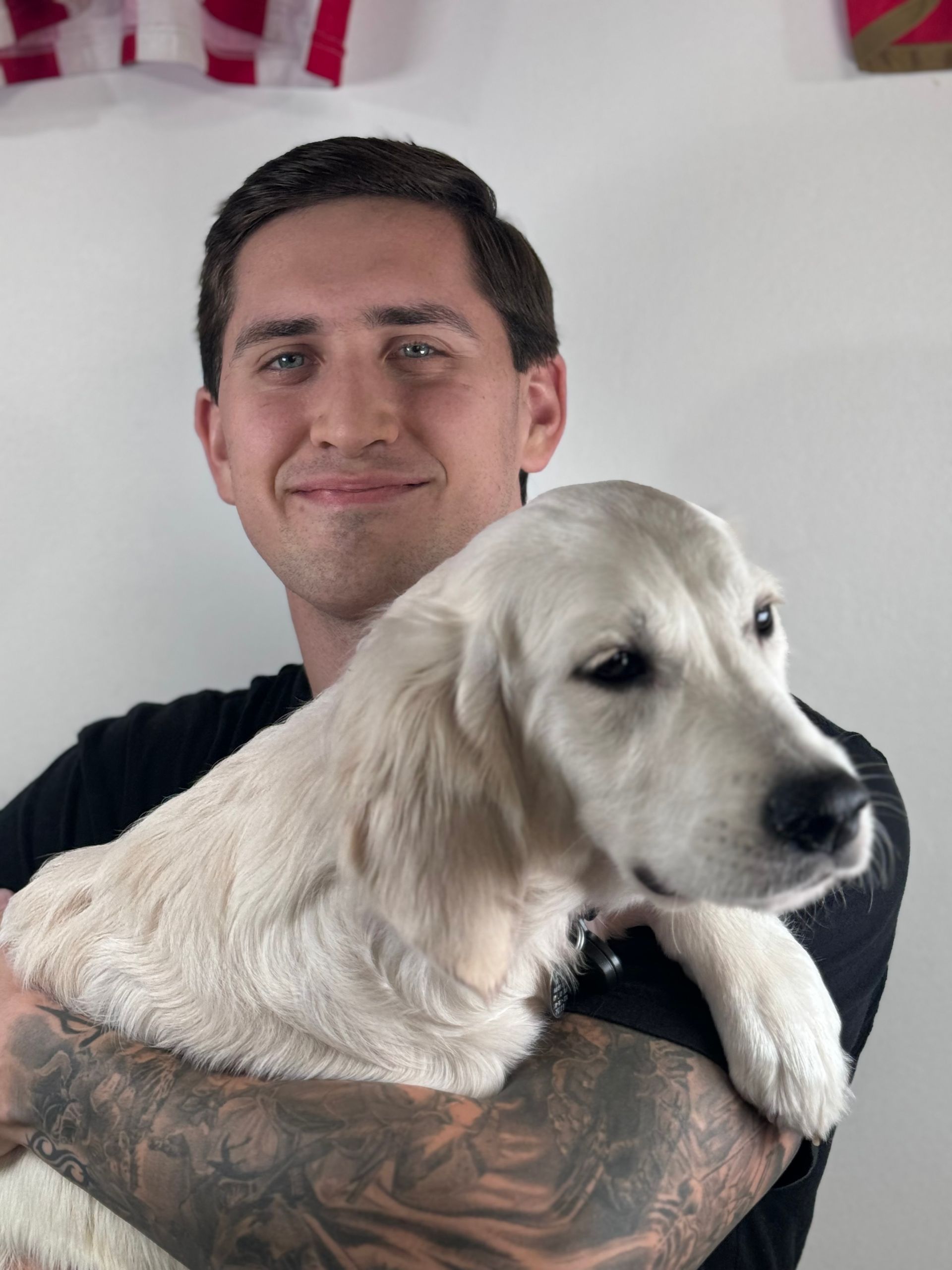 Man with tattoos holding a cream-colored dog, both smiling in front of a white background.