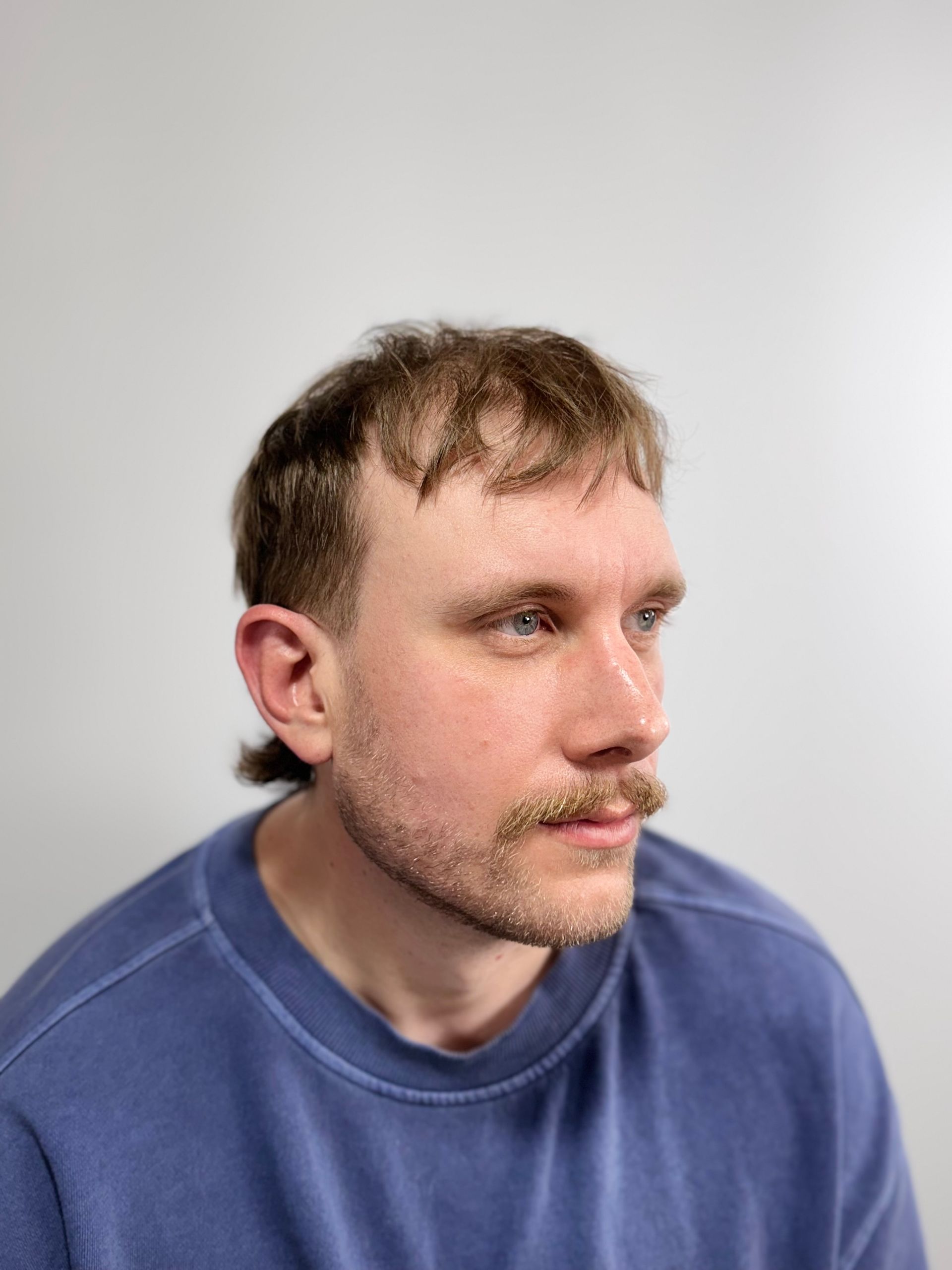 Man with light skin and dark hair looking right, wearing blue shirt, small mustache.