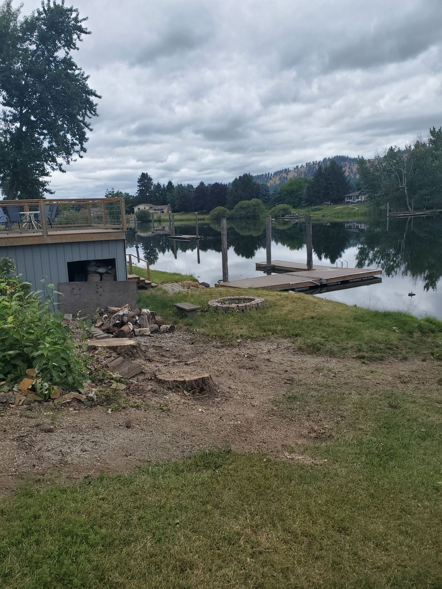 Lake view with a wooden dock, gray shed, and grassy shoreline under a cloudy sky.