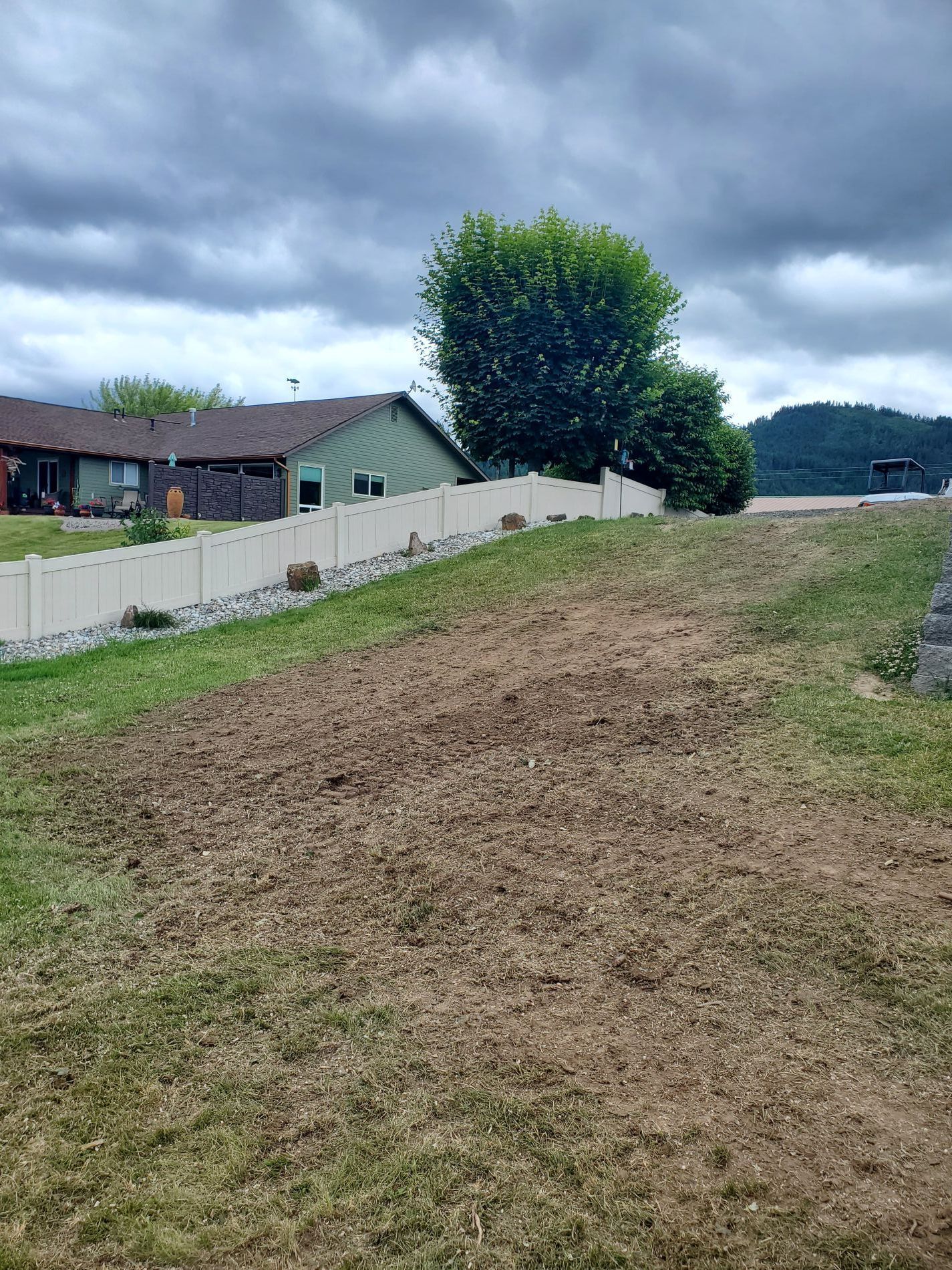 A yard with bare earth in the foreground, a white fence, and houses under a cloudy sky.