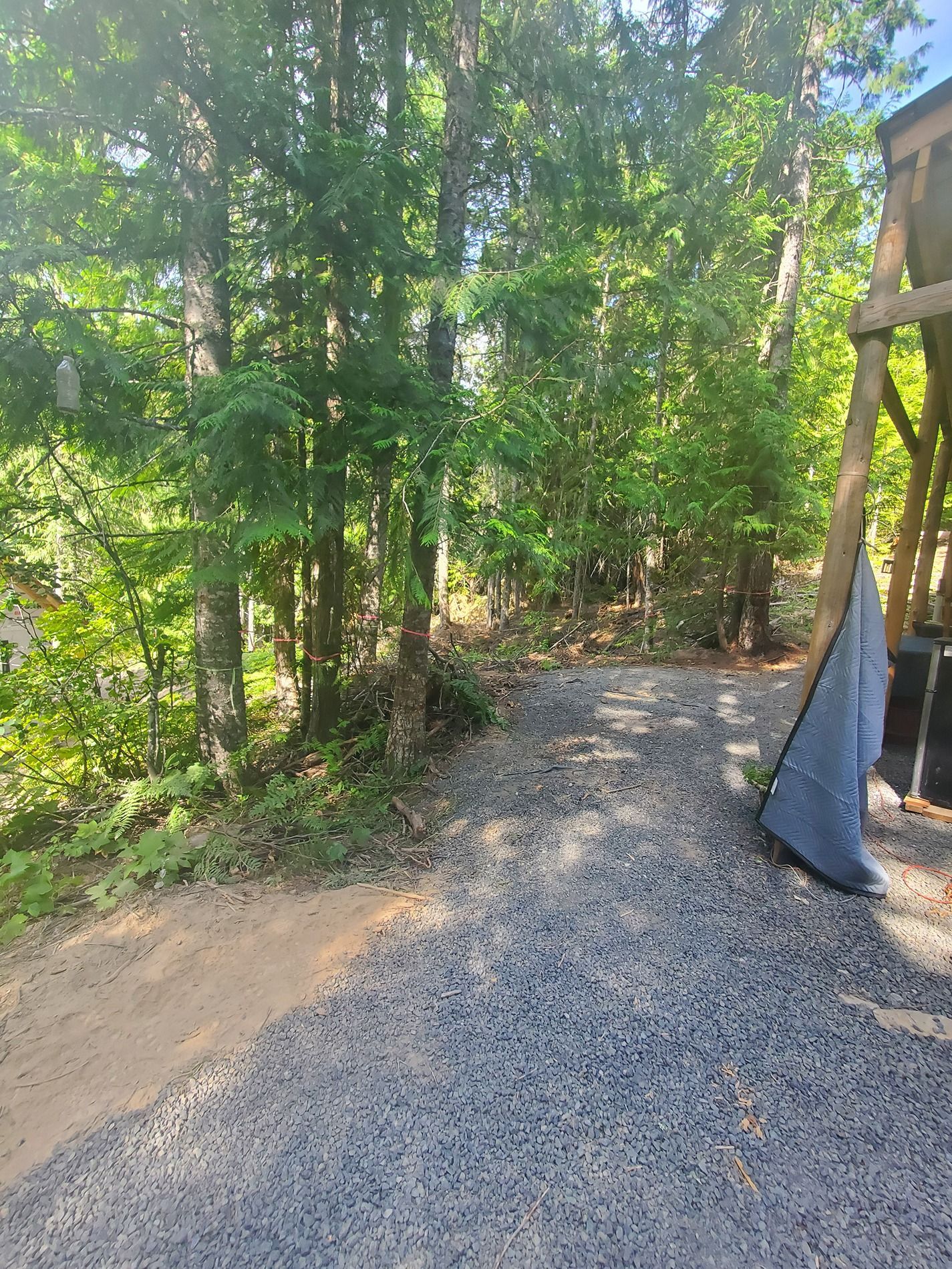 Gravel path leading into woods, with trees on either side and part of a wooden structure on the right.