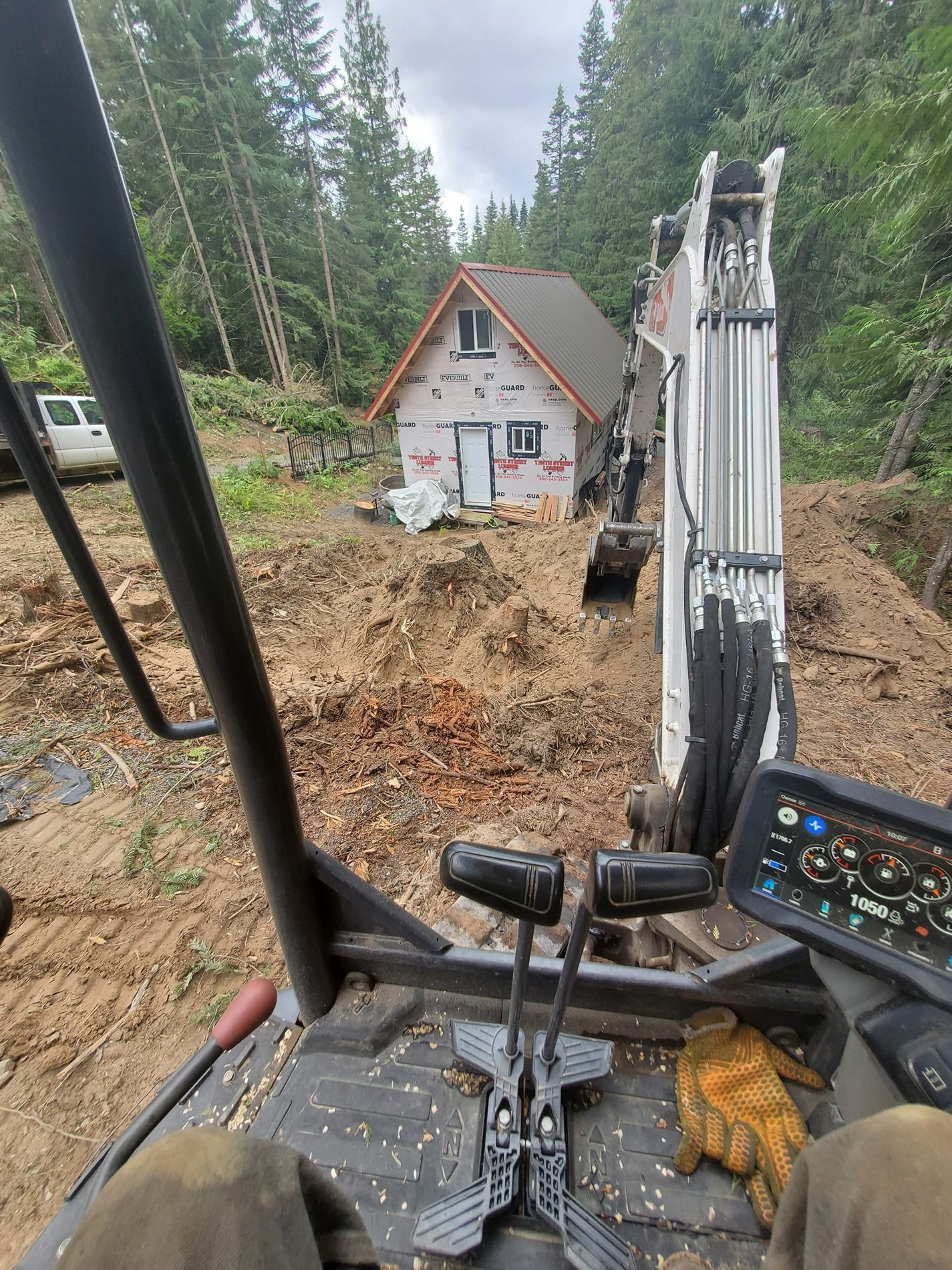 Excavator digging near a small house in a wooded area, seen from the operator's perspective.