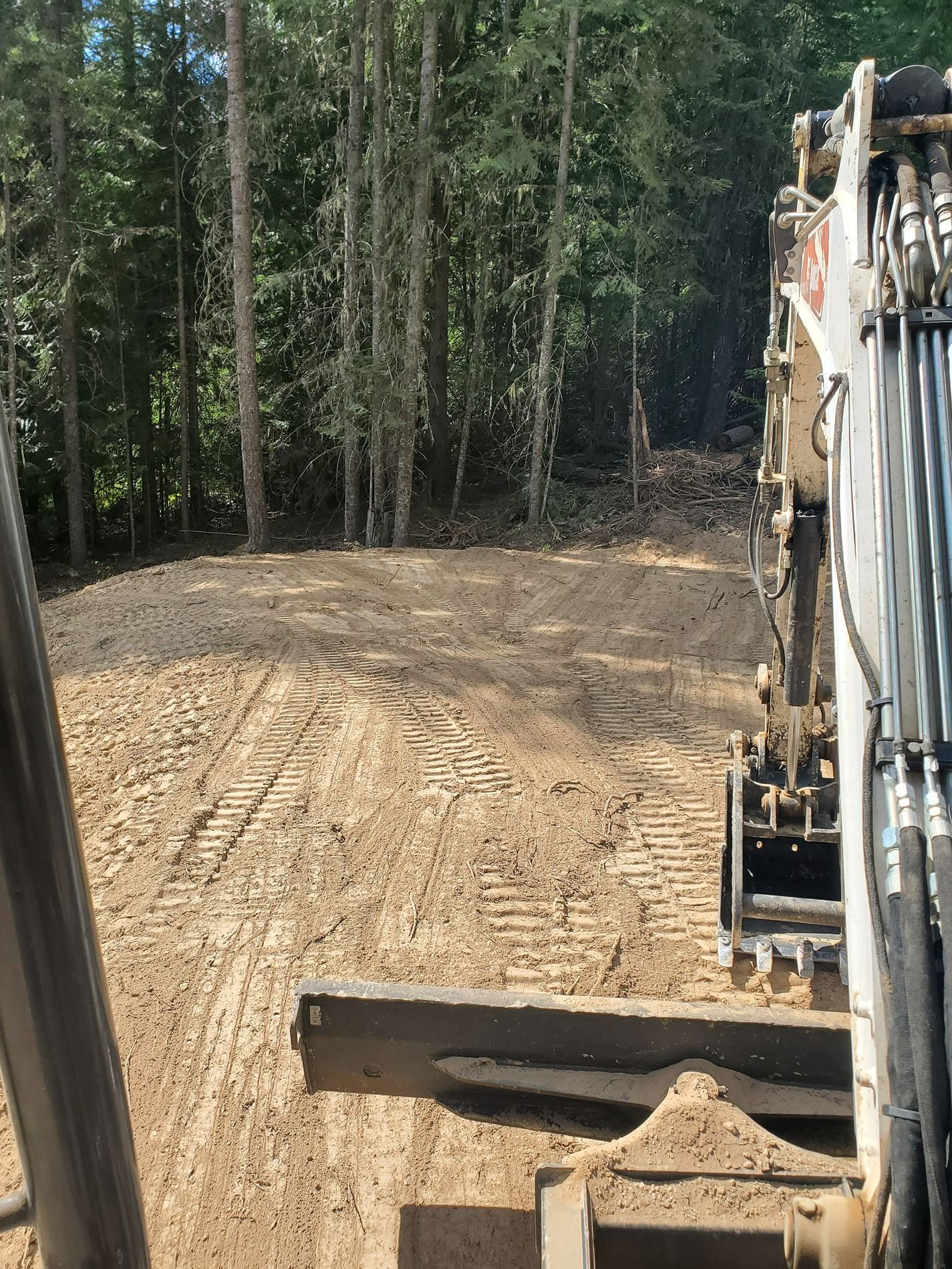 Excavator clearing land near a forest; the machine's arm and bucket are visible. Brown soil, trees in background.